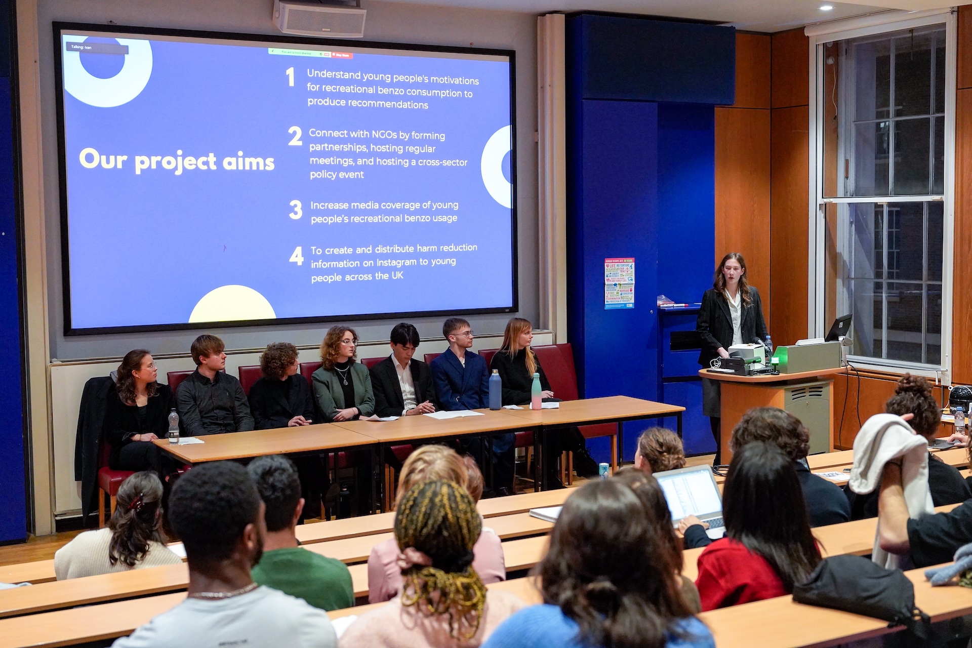 Seven volunteer members of the Benzo Research Project wearing smart attire sat in a line underneath a projector screen in a lecture theatre. Our Founder stood to the right at the podium presenting to the audience about the project's aims, as shown on the screen.